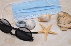 Sonnenbrille, Muscheln und medizinische Maske an Sandstrand.